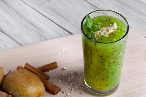 Close-up picture of a glass of beautiful light green, healthful smoothie with aromatic mint, spicy cinnamon and little nuts on a light desk background. Brown kiwi and cinnamon sticks next to a mix.