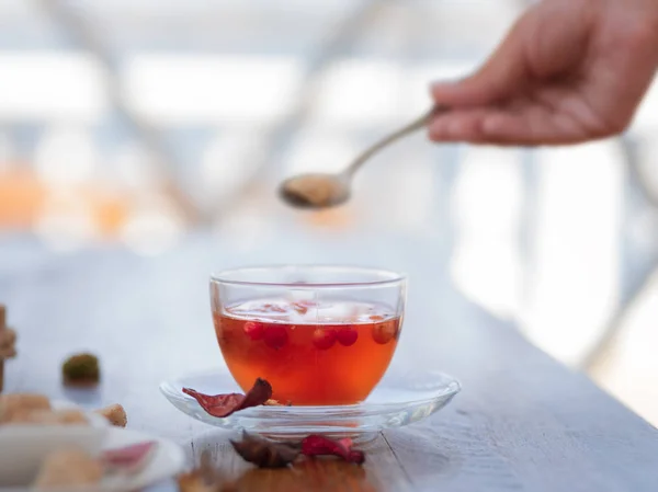 Close-up of a woman\'s hand with a spoon of sugar and a full teacup. Red tea in a cup with a spoon on a on a light blurred background. Tea with berries and leaves for a cafe breakfast. Copy space.