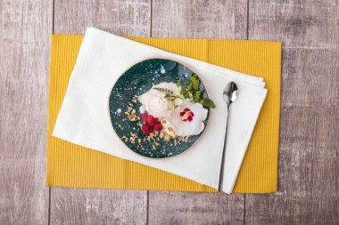 Vanilyalı dondurma mavi bir kabın içinde bir görünüm yukarıdan. Parlak sarı kumaş bir ahşap masa arka plan üzerinde beyaz dondurma. Pembe ahududu, fındık, yeşil nane ve bir çiçek ile soğuk bir aperatif.