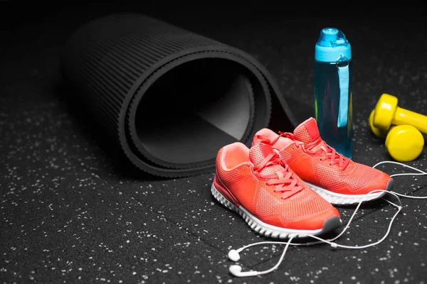 A composition of bright pink sports shoes, yellow dumbbells, blue sportive bottle and pilates mat on a black background. A colorful composition of sportive accessories. Healthy lifestyle.