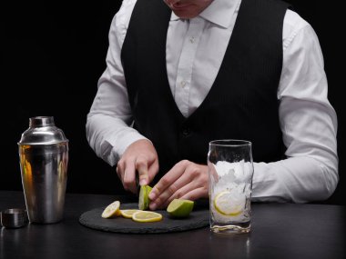 Meyveli bir alkollü içecek yapma siyah beyaz elbiseli şık bir barmen. Bir barmen kesme limes ve limon yanında bir bardak buz ve bir shaker bir bar counter üzerinde siyah bir arka plan.