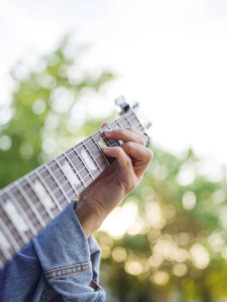 Pohled shora na mužské ruce hraje na kytaru v džínové bundy v parku na rozostřeného pozadí. — Stock fotografie