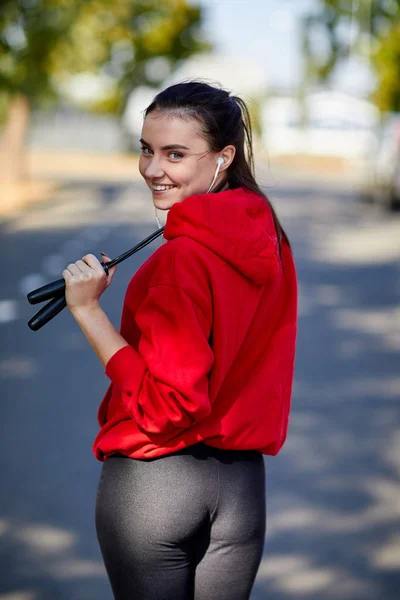 Sportif genç bir kadın, kulakları kulaklık ve bir atlama ipi onun elinde kırmızı bir kazak. — Stok fotoğraf