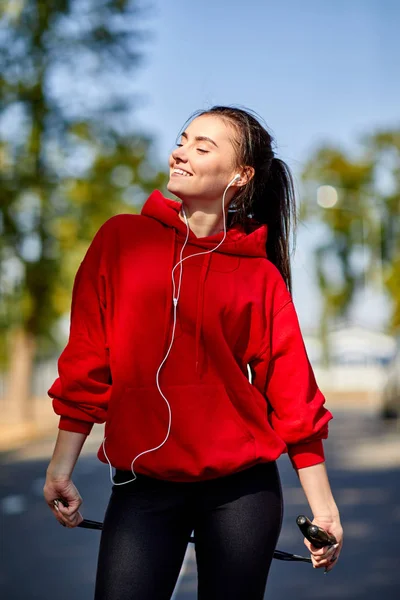Sportif genç bir kadın, kulakları kulaklık ve bir atlama ipi onun elinde kırmızı bir kazak. — Stok fotoğraf