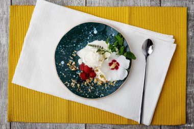 Vanilyalı dondurma mavi bir kabın içinde bir görünüm yukarıdan. Parlak sarı kumaş bir ahşap masa arka plan üzerinde beyaz dondurma. Pembe ahududu, fındık, yeşil nane ve bir çiçek ile soğuk bir aperatif.