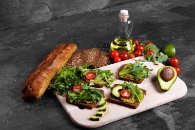 Besleyici avokado tost domates, baharatlı sos ve roka ahşap kesme masasında. Bir taş tablo arka plan üzerinde siyah ekmek, zeytin yağı, yeşil salata ve sebze dilimlenmiş. Kopya alanı.