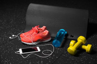 Parlak pembe spor ayakkabı, sarı dambıl, mavi sportif şişe ve siyah pilates mat siyah bir zemin üzerine yakın çekim bir resim. Sportif aksesuarlar renkli bir bileşimi. Sağlıklı yaşam tarzı.