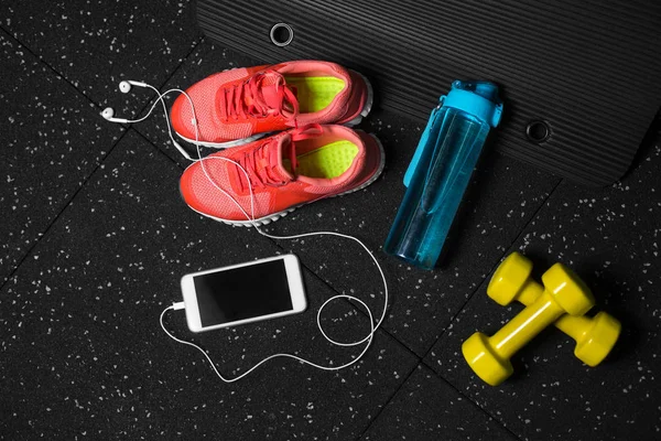 A view from above of bright pink training shoes, small yellow dumbbells, blue bottle for water and white smartphone with headphones on a black background. Comfortable female sports equipment.