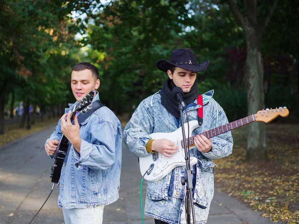 Happy young guys sings songs and plays guitar on a jeans jacket in a park on a natural background.