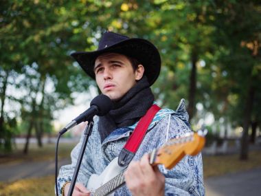 Genç adam şarkı söylüyor ve doğal bir arka plan üzerinde bir parkta bir kot ceket gitar çalar.