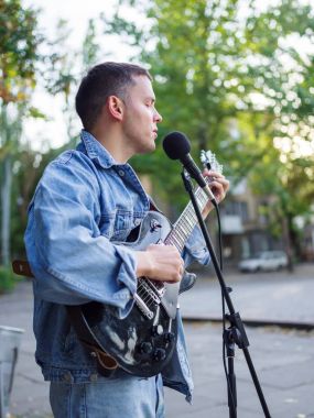 Genç adam şarkı söylüyor ve doğal bir arka plan üzerinde bir parkta bir kot ceket gitar çalar.