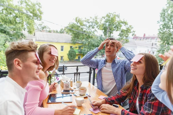 Teen friends having fun playing with stickers on foreheads on a cafe background. Friends activity concept. — Stock Photo, Image
