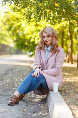 Beautiful blonde is sitting on the curb in the park with coffee. clipart