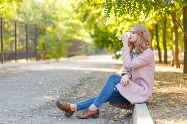 Красива блондинка сидить на бордюрі в парку з кавою . — стокове фото