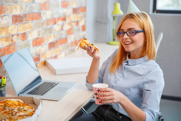 Una chica comiendo pizza en la oficina delante de un portátil —  Fotos de Stock