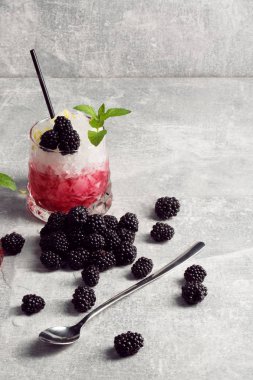 Bir bardak taze kırmızı meyve kokteyli ve buz, böğürtlen ve yeşil sappe nane, parlak gümüş çay kaşığı gri hafif bir arka plan üzerinde bırakır.