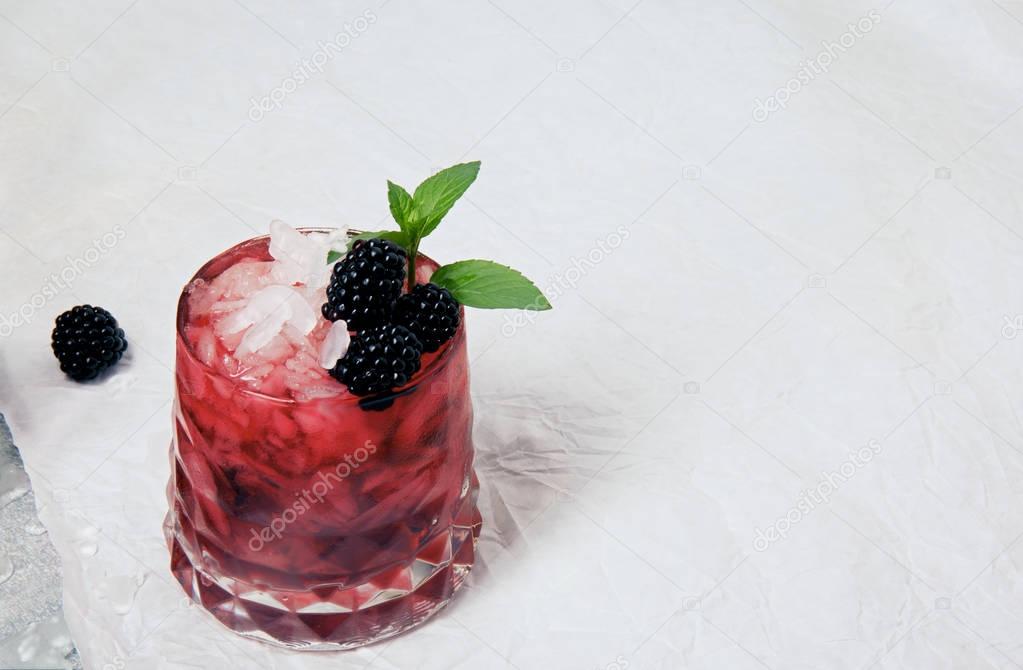 Summer coctail with ice cubes, fresh green mint and sappy blackberries in a pink glass on a white background, copy space, blackberry on a white table-cloth.