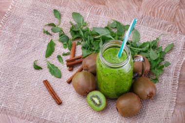 Ahşap arka plan üzerinde renkli bir saman bir mason kavanoza bir sağlıklı kivi güler yüzlü bir manzara yukarıdan. Egzotik bütün ve kesme kivi, tarçın sopa ve bir içecek beyaz bir bez üzerinde bırakır.