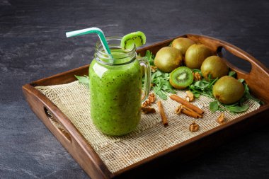 Koyu gri arka plan üzerinde soğuk kivi içecek close-up. Renkli bir saman bir mason kavanoza bir kokteyl. Renkli kivi, tarçın sopa ve yanındaki bir ahşap tepsi içinde soğuk bir içecek nane yaprakları. 