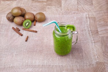 Bir mason kavanoza bir dilim beyaz peçete üzerinde yeşil taze kivi duran ve çizgili içme ile kivi güler yüzlü saman dolu. Tarçın ve kivi hafif ahşap arka plan üzerinde. Kopya alanı.