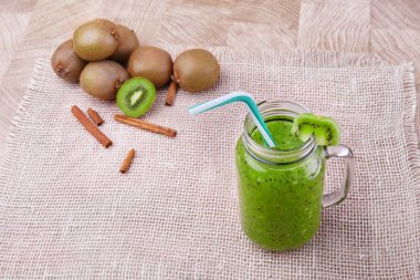 Bir mason kavanoza bir dilim beyaz peçete üzerinde yeşil taze kivi duran ve çizgili içme ile kivi güler yüzlü saman dolu. Tarçın ve kivi hafif ahşap arka plan üzerinde. Kopya alanı.