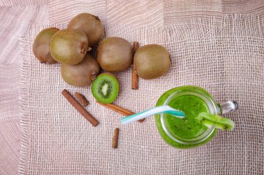 Kivi güler yüzlü çizgili saman ve hafif kahverengi ahşap arka plan üzerinde tarçınlı beyaz peçeteye yeşil taze kivi ve kivi ahşap bir masa üzerinde bir dilim ile tam bir mason kavanoza Üstten Görünüm.