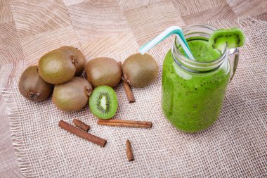 Bir mason kavanoza bir dilim beyaz peçete üzerinde yeşil taze kivi duran ve çizgili içme ile kivi güler yüzlü saman dolu. Tarçın ve kivi hafif ahşap arka plan üzerinde. Kopya alanı.