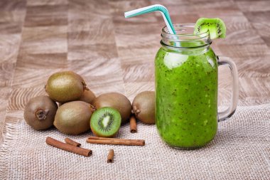 Bir mason kavanoza bir dilim beyaz peçete üzerinde yeşil taze kivi duran ve çizgili içme ile kivi güler yüzlü saman dolu. Tarçın ve kivi hafif ahşap arka plan üzerinde. Kopya alanı.