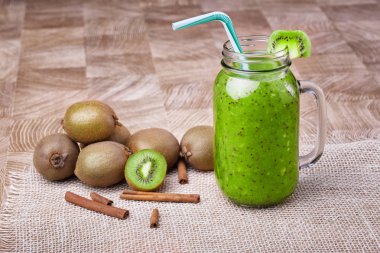 Bir mason kavanoza bir dilim beyaz peçete üzerinde yeşil taze kivi duran ve çizgili içme ile kivi güler yüzlü saman dolu. Tarçın ve kivi hafif ahşap arka plan üzerinde. Kopya alanı.