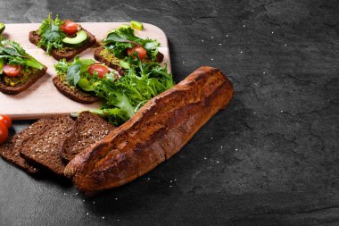 ?kaybetmek'lı Fotoğraflar rustik, taze pişmiş, gevrek ve gevrek bir siyah tablo arka plan üzerinde siyah baget ekmek dilimlenmiş. Ekmek sebze sandviç ve yeşil roka yaprakları ile kesim masası yanında.