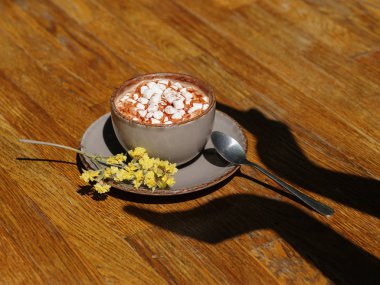 Bir gri kahve fincanı bir close-up cappuccino ve beyaz marshmallow ile dolu. Ahşap masa arka plan üzerinde tatlı lezzetli içecek. Elinde bir fincan çiçek yanında bir gölge ile. Kopya alanı.
