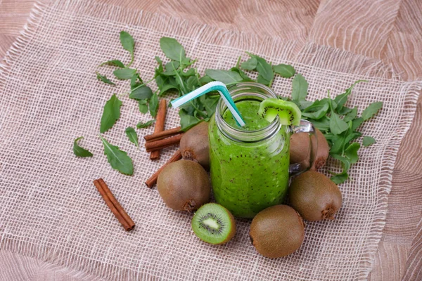 A top view of a healthy kiwi smoothie in a mason jar with a colorful straw and slice of fruit on a top on a wooden background. Exotic whole and cut kiwi, cinnamon sticks and leaves on a white cloth.