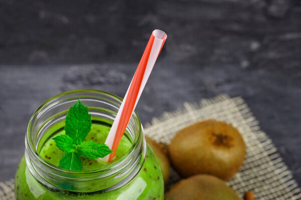 A close-up of a jar filled with a sturated green smoothie with a striped straw and fresh mint leaves. A healthy, refreshing and exotic cocktail and whole kiwis on a black table background.  