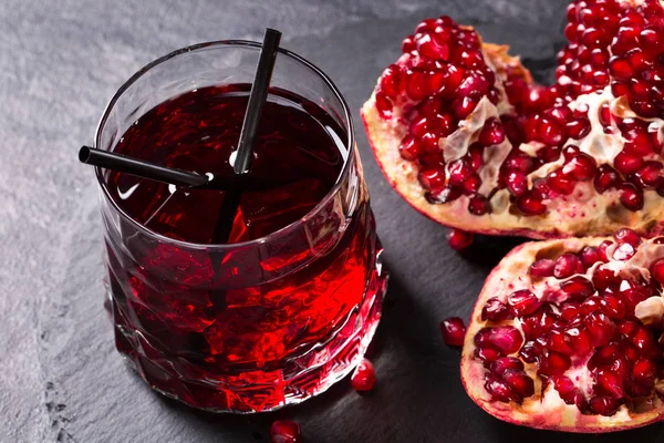 A big transparent glass of pomegranate juice with two straws on a dark gray background. A ripe garnet broken into pieces on a table. The organic red beverage with exotic fruit. Healthful ingredients.