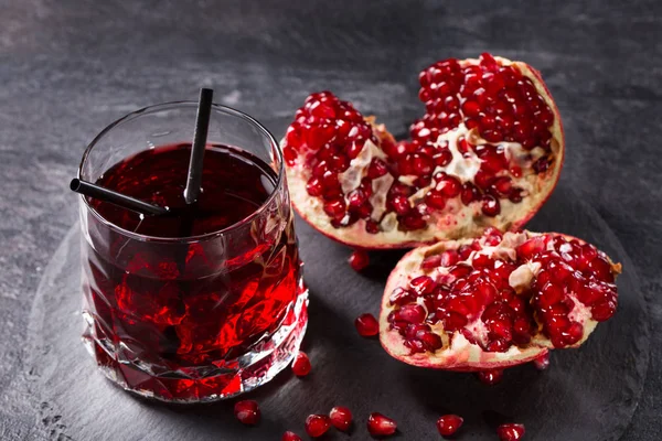 A big transparent glass of pomegranate juice with two straws on a dark gray background. A ripe garnet broken into pieces on a table. The organic red beverage with exotic fruit. Healthful ingredients.