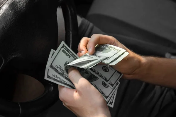 Strong man's hands counting paper money in a new automobile on a blurred black background. A successful male holding hundreds of dollars in cash. Salary, earning, payment, payout concept.
