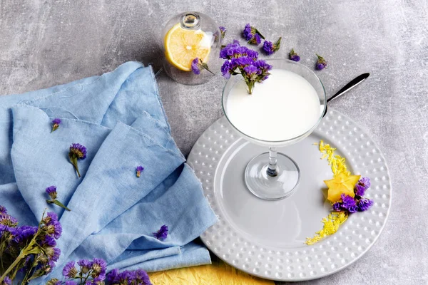 A beautiful composition of a margarita glass full of white milkshake and colorful fabric on a gray table background. Organic, fresh, healthful and cold milk smoothie with purple flowers.