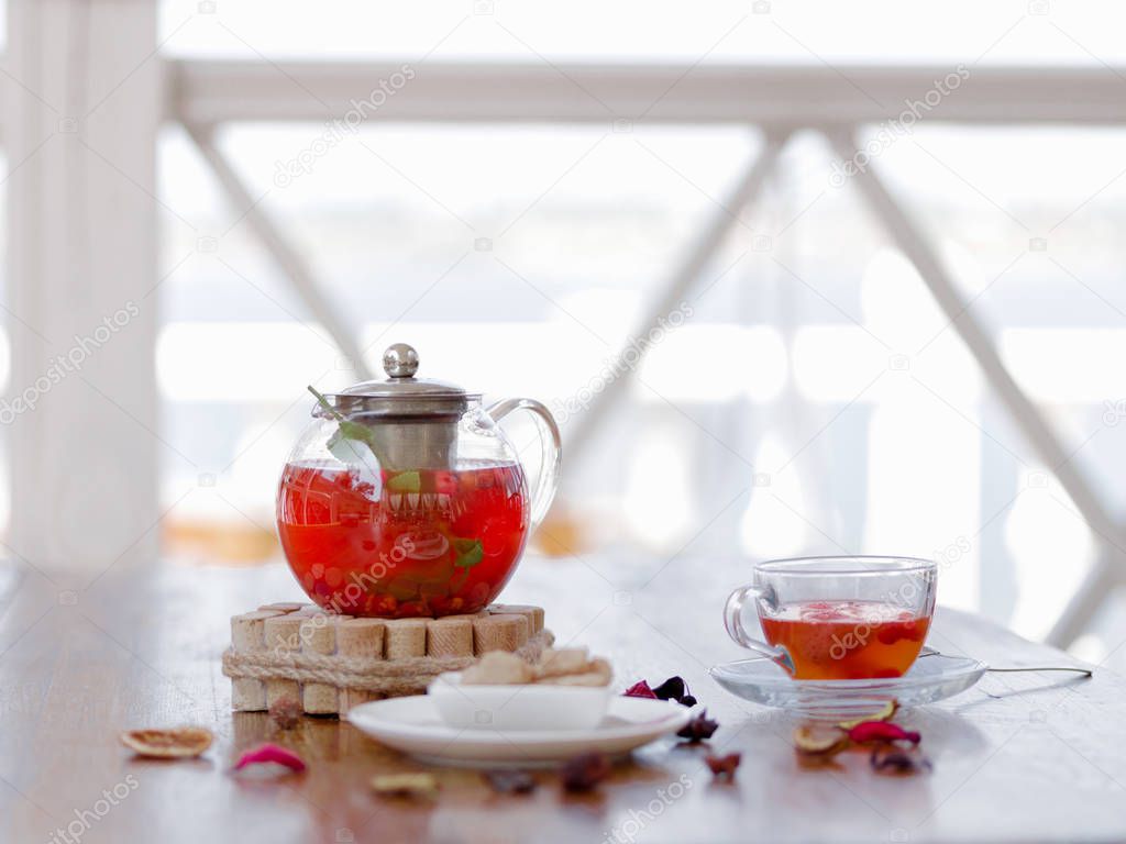 A tender composition of a transparent teapot full of strawberry tea with red currant on a wooden table in a restaurant. A berries red beverage with spices on a blurred background. Organic drink.