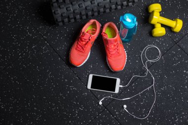 Mavi sportif şişe, turuncu Eğitim ayakkabı, sarı salak-bells, siyah pilates mat ve modern telefon kulaklık siyah bir arka plan ile bir görünüm yukarıdan. Eğitim, konsept spor. Kopya alanı.