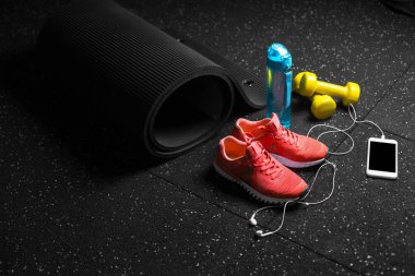 Sportif aksesuarlar, spor eğitim siyah zemin arka plan üzerinde renkli bir bileşimi. Mavi sportif şişe, parlak turuncu spor ayakkabı, sarı salak-bells, siyah pilates mat. Eğitim kavramı.