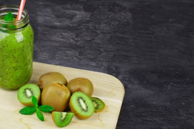 Tam bir mason kavanoza çizgili saman ile kivi içecek, taze kivi ve yeşil ışık ahşap tahtası koyu bir taş zemin üzerine nane bırakır. Yaz meyve. Kopya alanı.