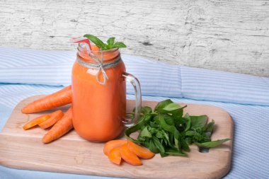 Bir mason kavanoza bir detoks içecek tam bir yakın çekim resim. Mavi kumaş arka plan üzerinde serinletici bir havuç yüzlü. Olgun havuç tüpler ve yeşil fesleğen yaprakları kesme masanın yanında bir vitamin içeceği.