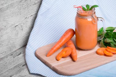 Mavi kumaş arka plan üzerinde serinletici bir havuç yüzlü bir yakın çekim resim. Bir mason kavanoza bir detoks içecek dolu. Olgun havuç tüpler ve yeşil fesleğen yaprakları kesme masanın yanında bir vitamin içeceği.