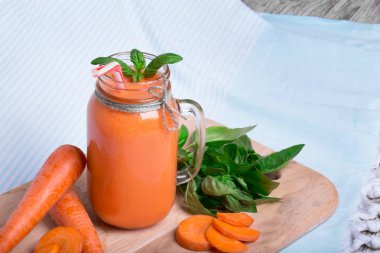 Bir vitamin içeceği olgun havuç tüpler ve yeşil fesleğen yaprakları kesme masanın yanında bir makro resim. Mavi kumaş arka plan üzerinde serinletici havuç yüzlü. Bir detoks içecek tam bir mason kavanoz.