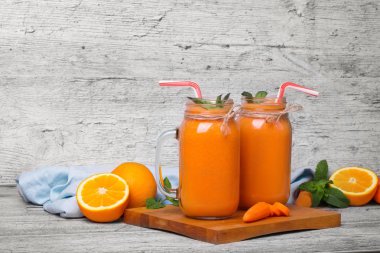 Şeffaf bir çift kesme masa üzerinde duran nane ile Besleyici, vegan turuncu sebze suyu dolu kavanozları. Kıyılmış turuncu meyveler ve rustik gri bir arka plan üzerinde soyulmuş havuç. Kopya alanı.