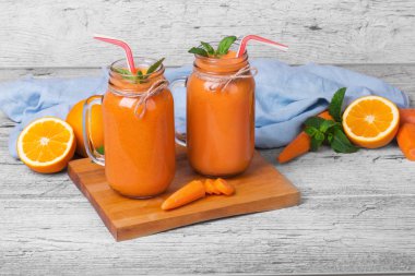 Şeffaf bir çift kesme masa üzerinde duran nane ile Besleyici, vegan turuncu sebze suyu dolu kavanozları. Kıyılmış turuncu meyveler ve rustik gri bir arka plan üzerinde soyulmuş havuç. Kopya alanı.