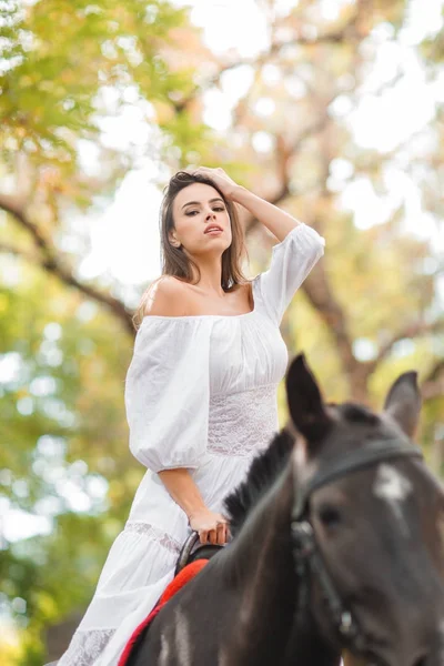 Ridning. Vacker ung kvinna i en vit klänning som rider på en brun häst utomhus. — Stockfoto