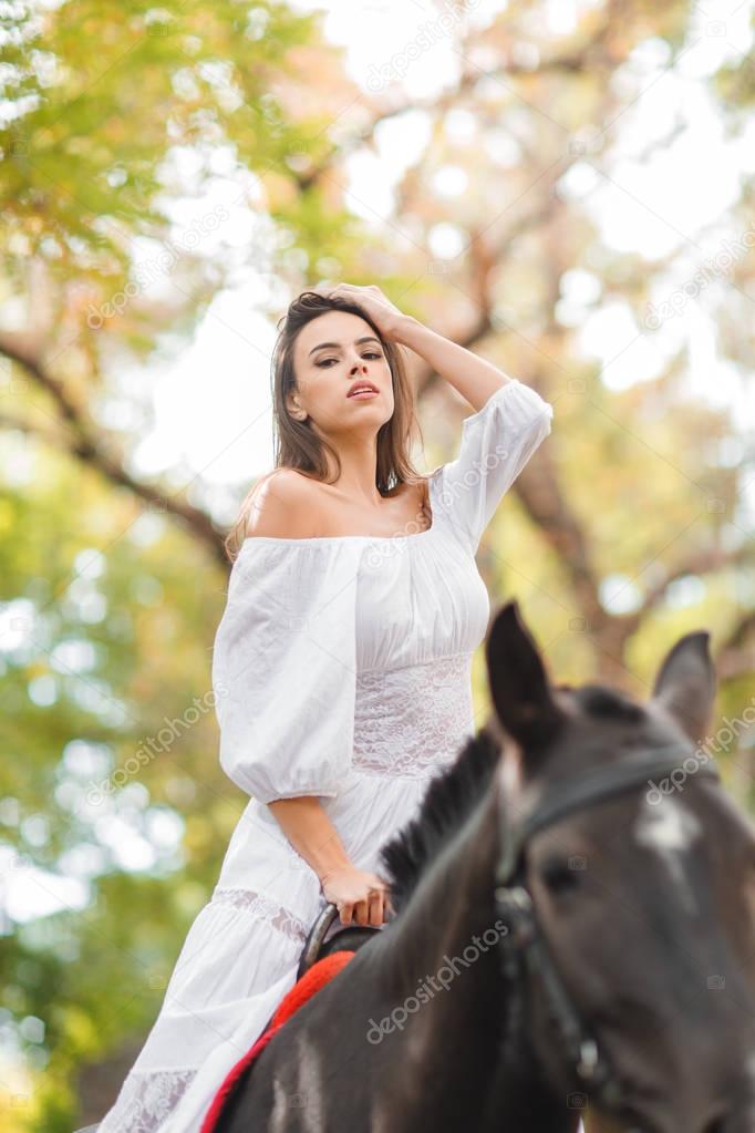 Horseback riding. Beautiful young woman in a white dress riding on a brown horse outdoors.