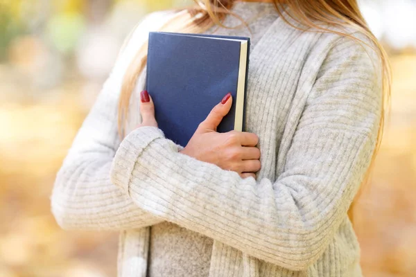 Uma menina bonita em uma camisola branca está segurando um livro e um tablet no fundo do parque de outono . — Fotografia de Stock