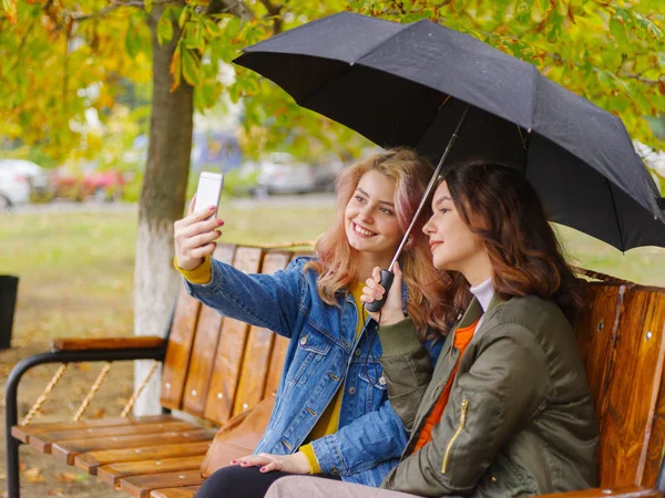 Girls do selfie under an umbrella on a bench — Stock Photo, Image